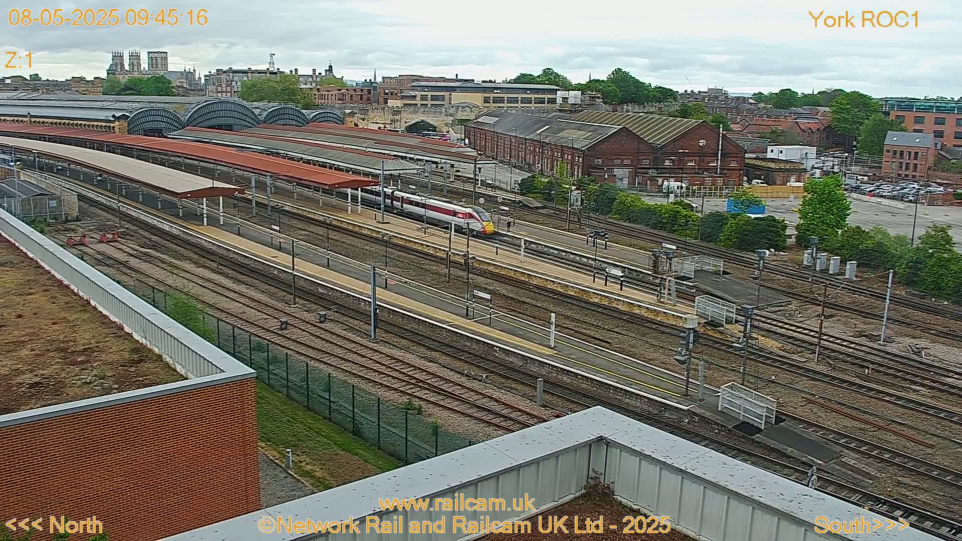 A high-angle view of a train station on an overcast day, showing a high-speed train arriving at multiple platforms surrounded by tracks and buildings.