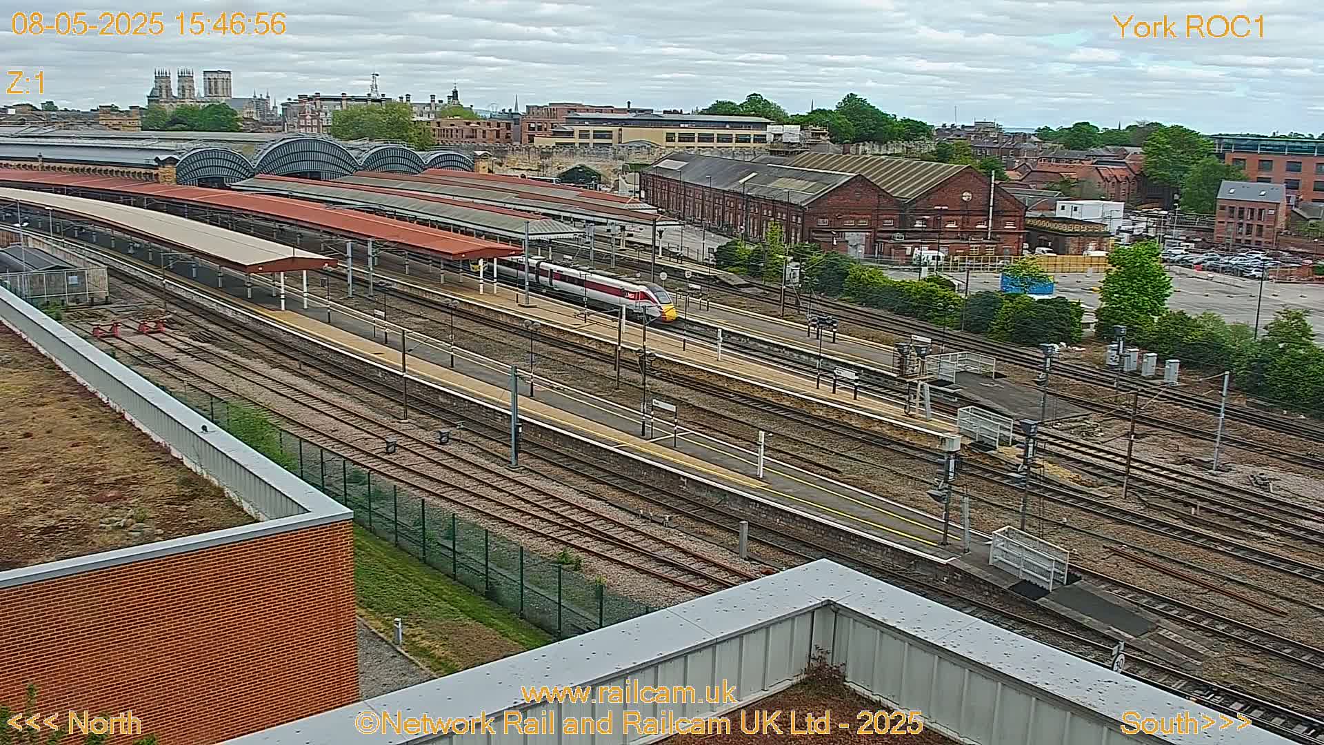 A high-angle view of a train station on an overcast day shows a high-speed train passing through numerous railway tracks and platforms, with a city and cathedral visible in the background.
