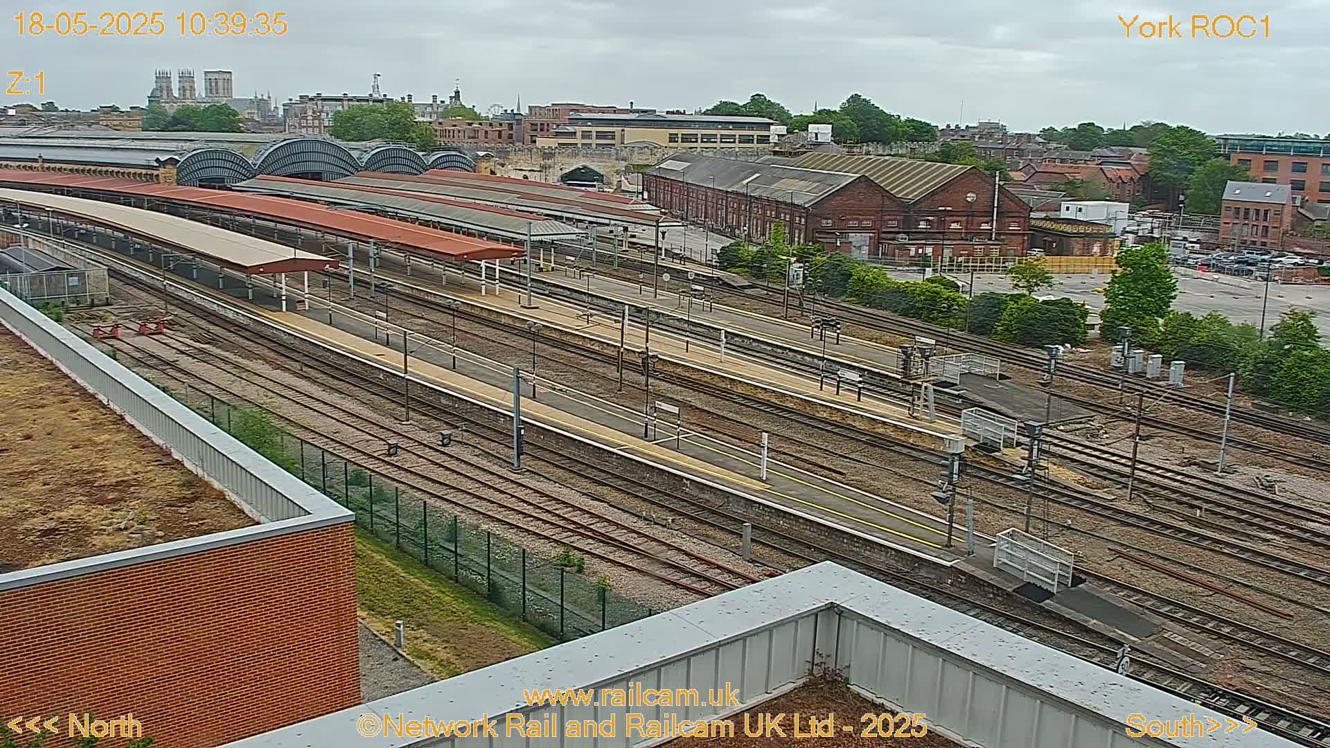 An overcast day reveals a high-angle view of a sprawling train station with numerous tracks and platforms, surrounded by buildings and greenery.