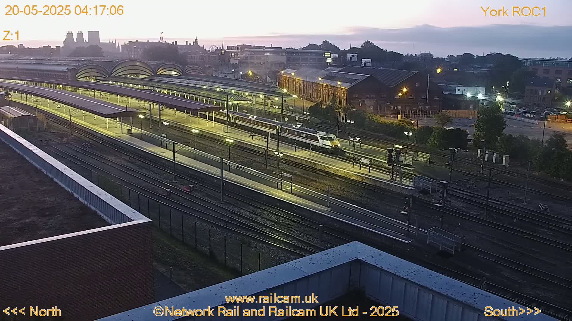 A high-angle, twilight view of a train station with a train passing through numerous tracks and platforms under a mostly clear sky.