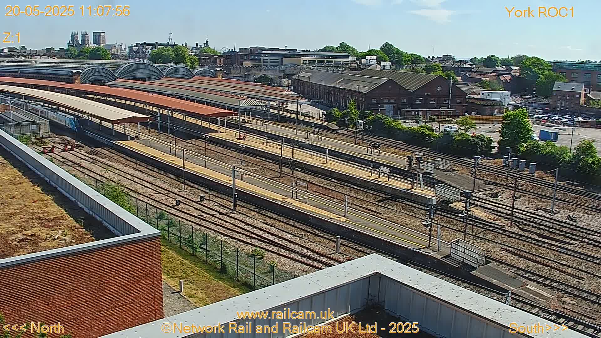 A high-angle view of a train station on a sunny day, showing multiple train tracks, platforms, and surrounding buildings.