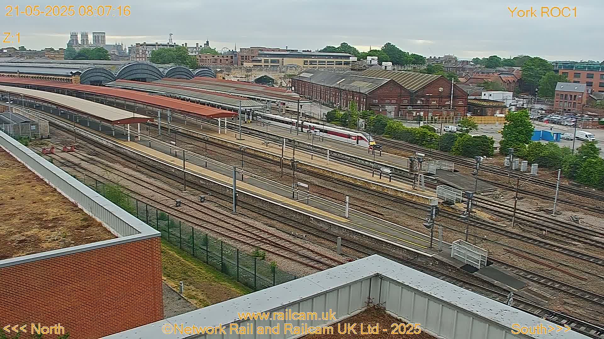 A high-angle view of a train station on an overcast day, showing numerous train tracks, platforms with covered waiting areas, and a high-speed train passing through.