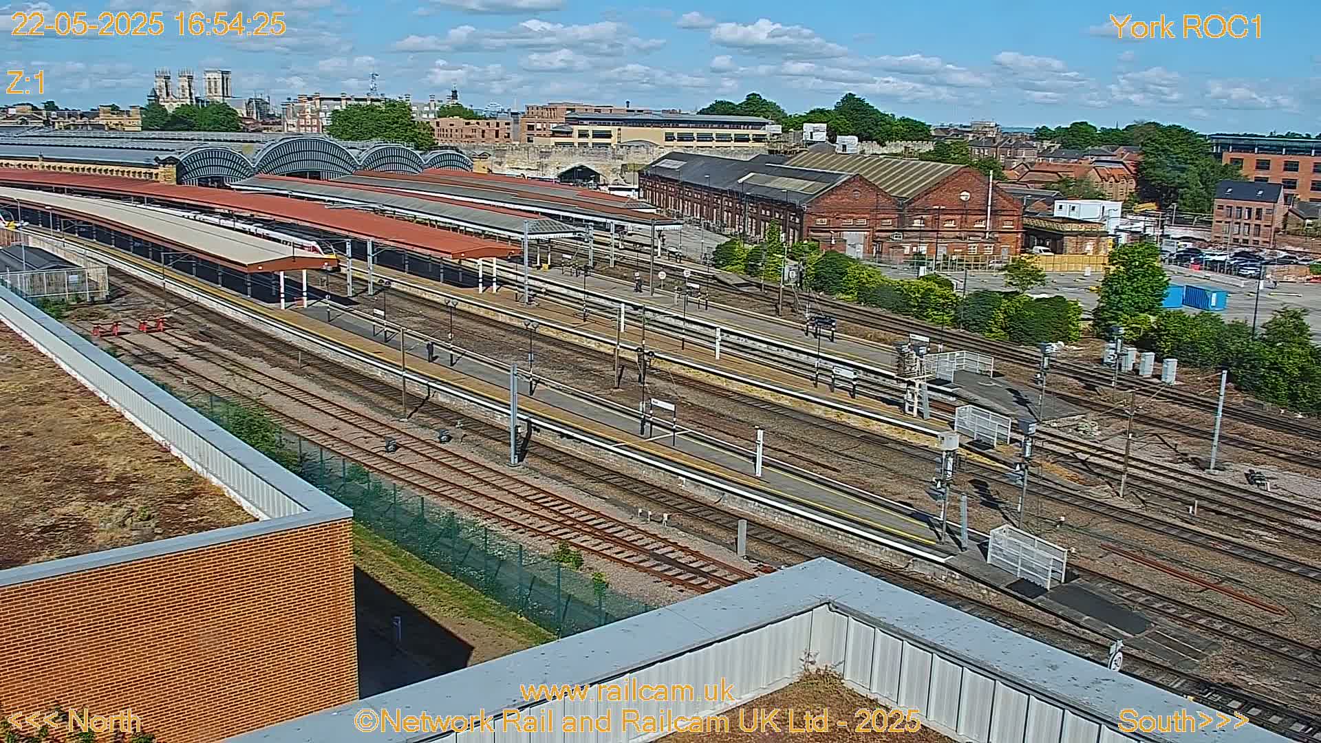 A high-angle view of a train station on a sunny day, showing numerous train tracks, platforms with trains, and surrounding buildings.
