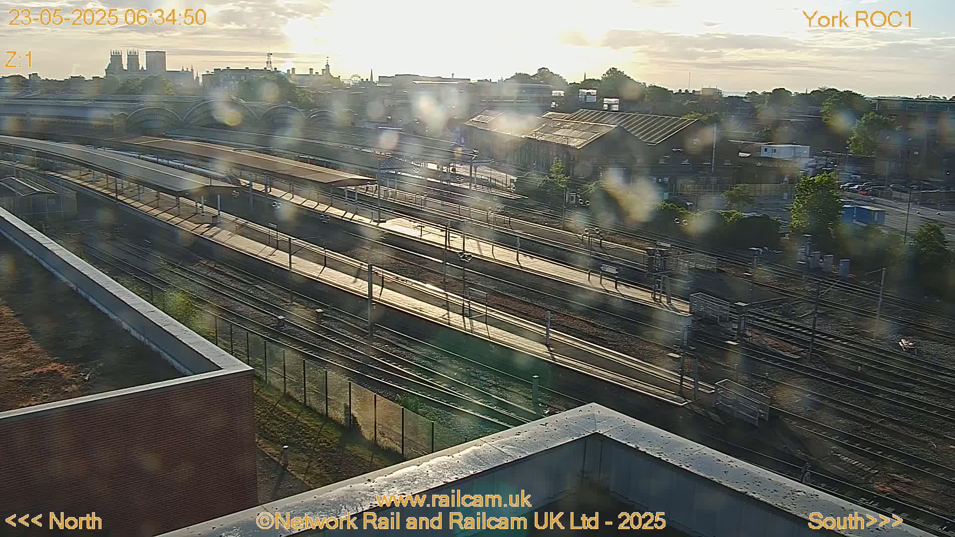 A high-angle, long shot of a railway station and its surrounding area on a partly sunny morning, with some lens glare.