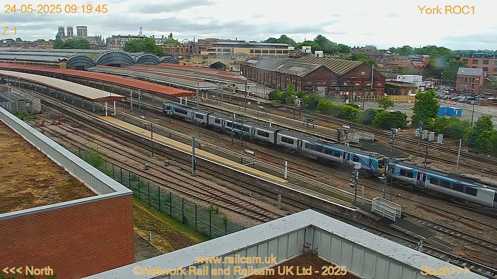 An elevated view of a train station on an overcast day shows multiple train tracks with two passenger trains at a platform, surrounded by industrial buildings and a distant city skyline.