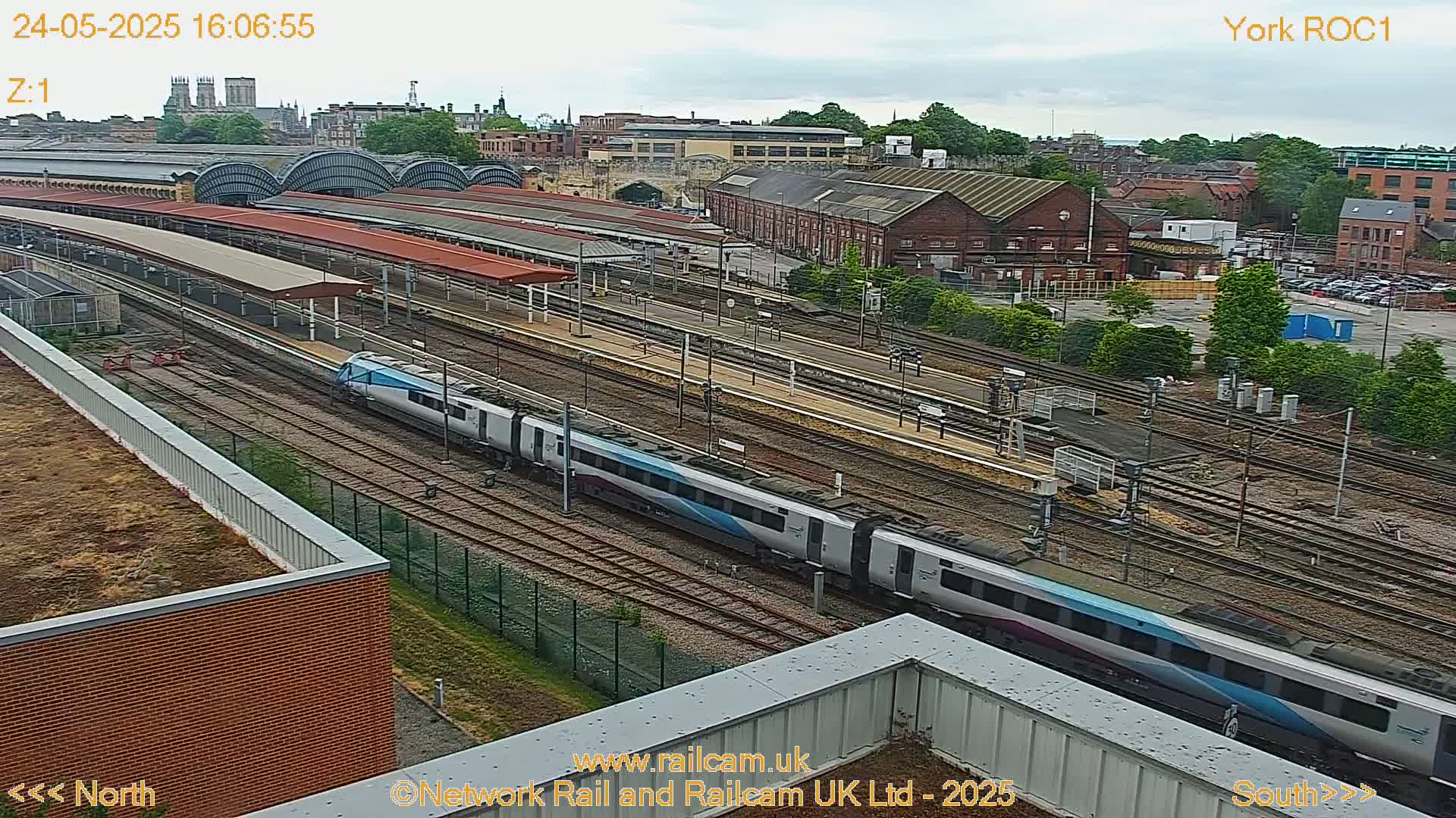 A long passenger train moves through a railway station on an overcast day, with a cityscape visible in the background.