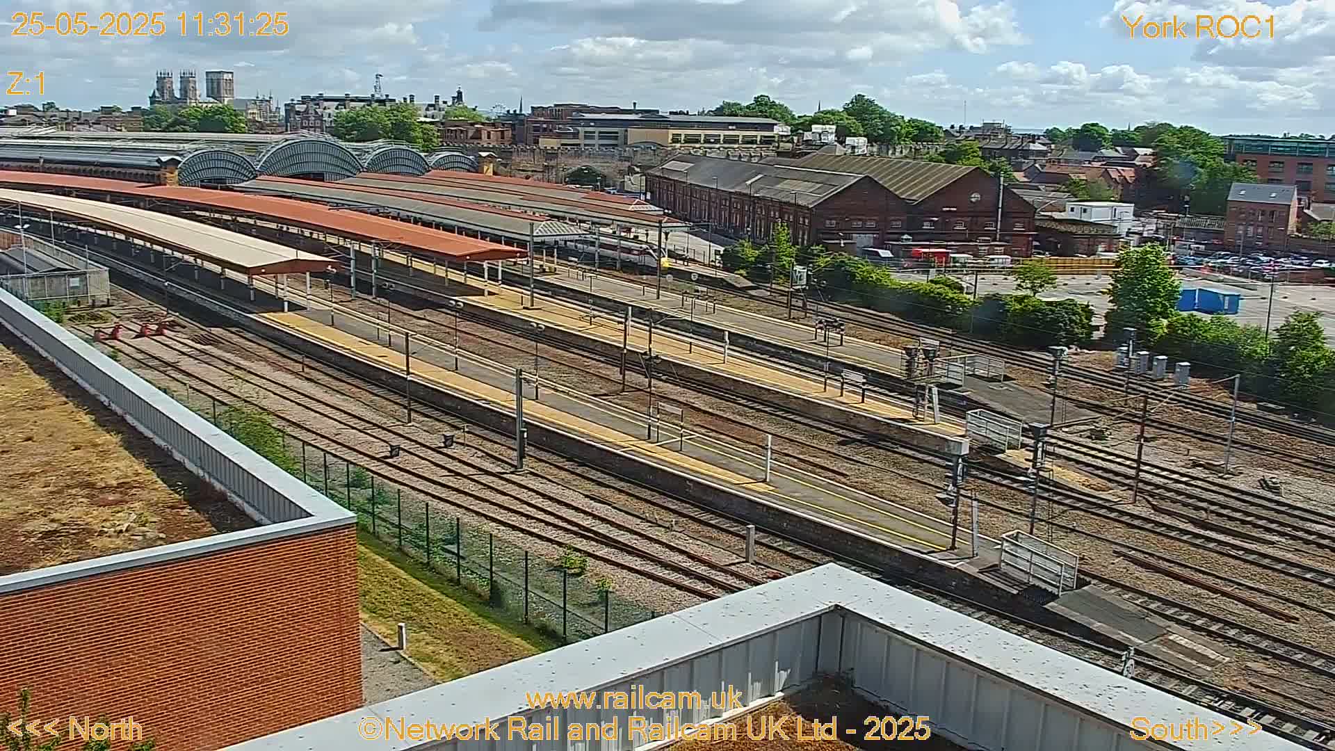 A high-angle view of a train station on a partly cloudy day, showing numerous train tracks and platforms, with a city skyline visible in the background.