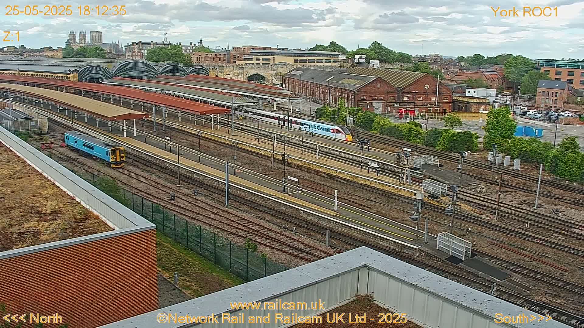 A high-angle view of a train station with multiple train tracks, platforms, and trains, under a partly cloudy sky.