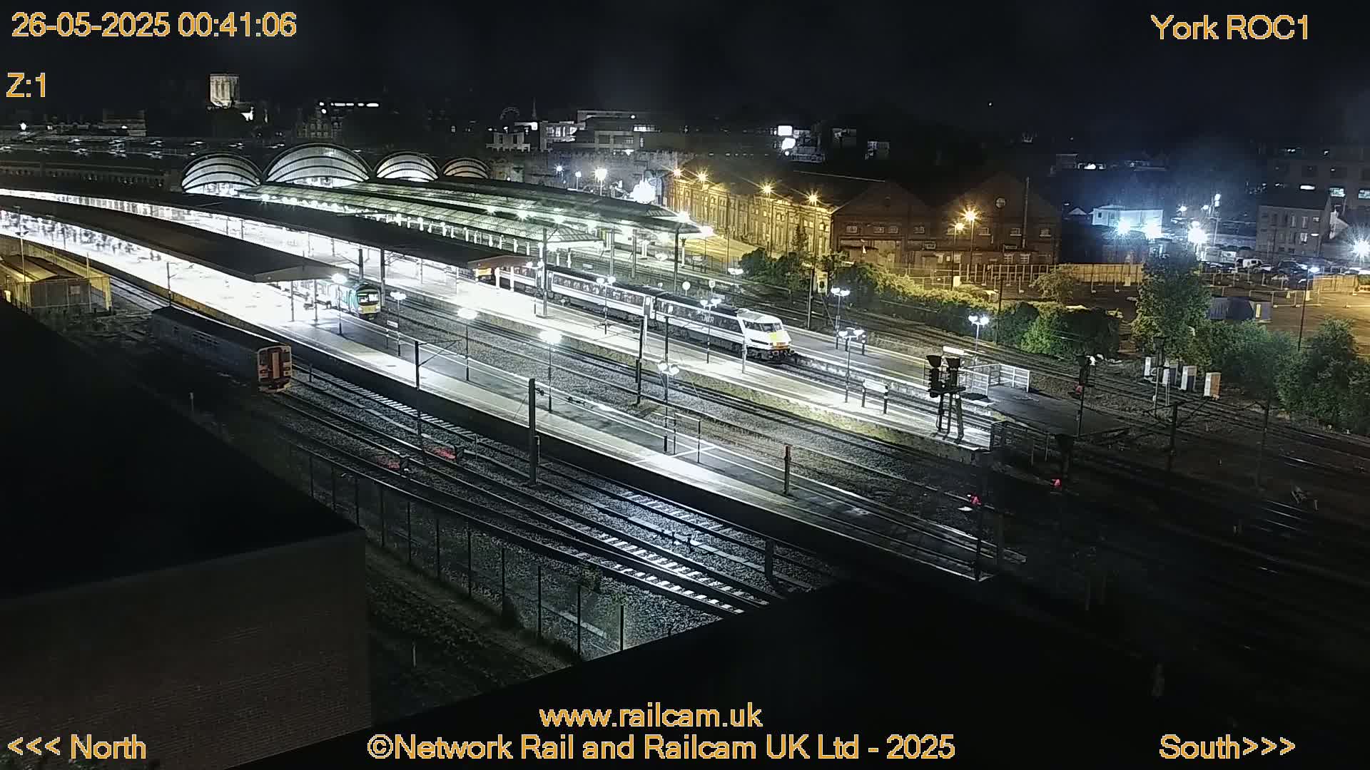 A nighttime aerial view of a train station with several trains on the tracks and platforms under covered areas, brightly lit against a dark background.