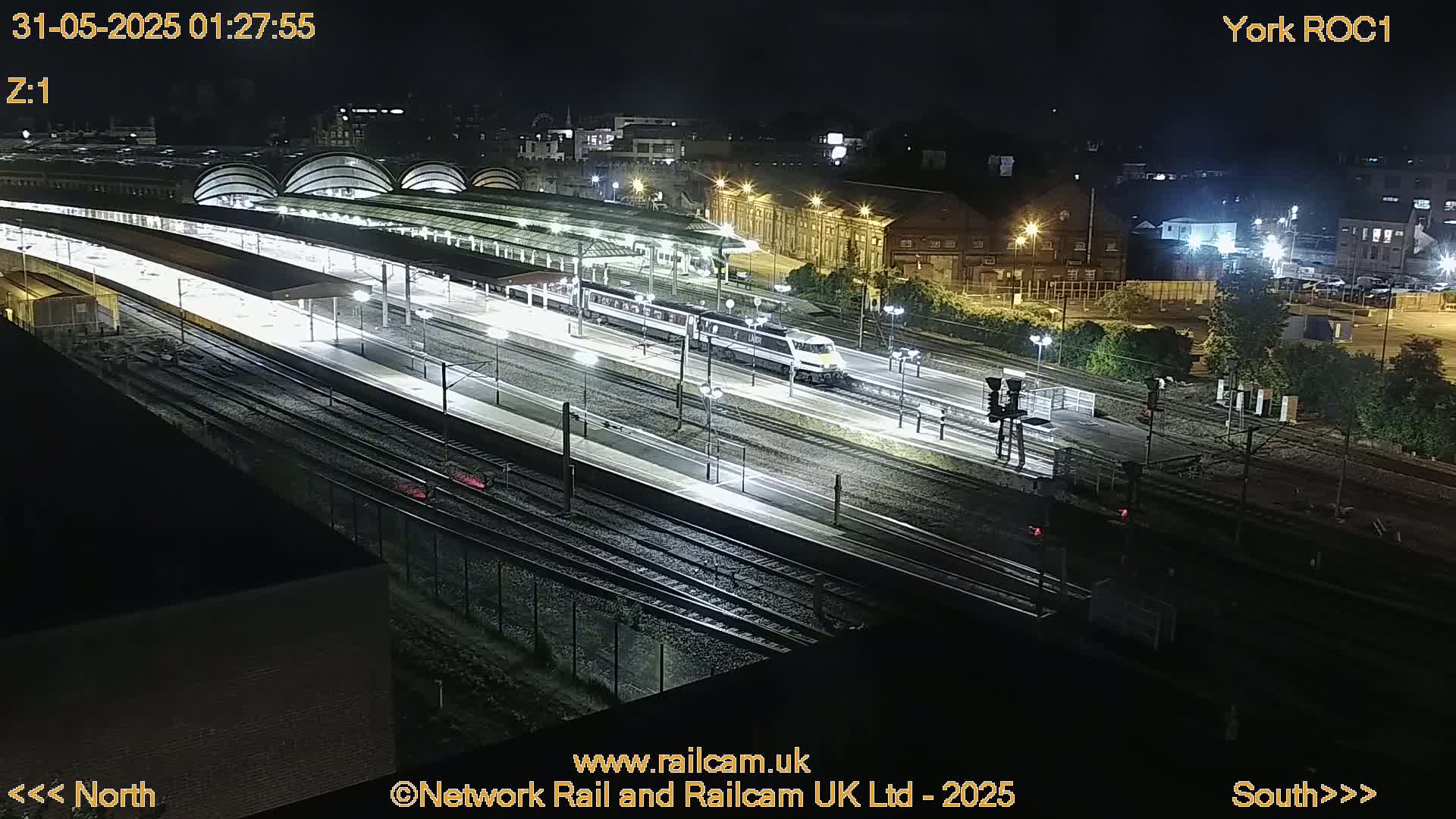 A long, brightly lit train travels through a railway station at night.