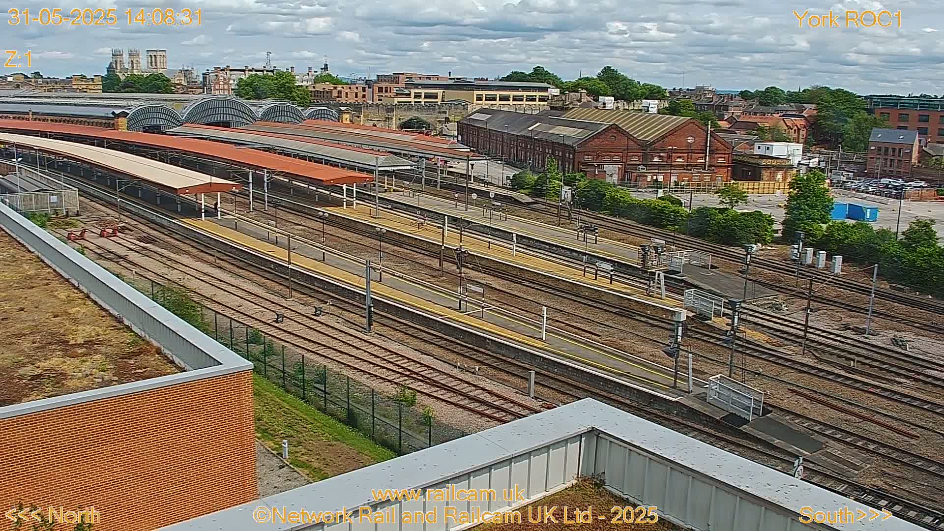 A high-angle view of a train station on a partly cloudy day, showing multiple train tracks, platforms with shelters, and surrounding buildings.