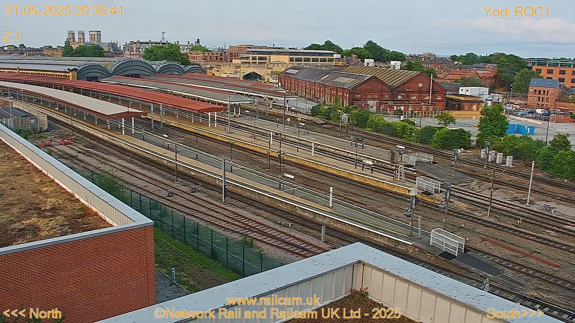 An elevated view of a railway station and surrounding urban area under a mostly clear evening sky.