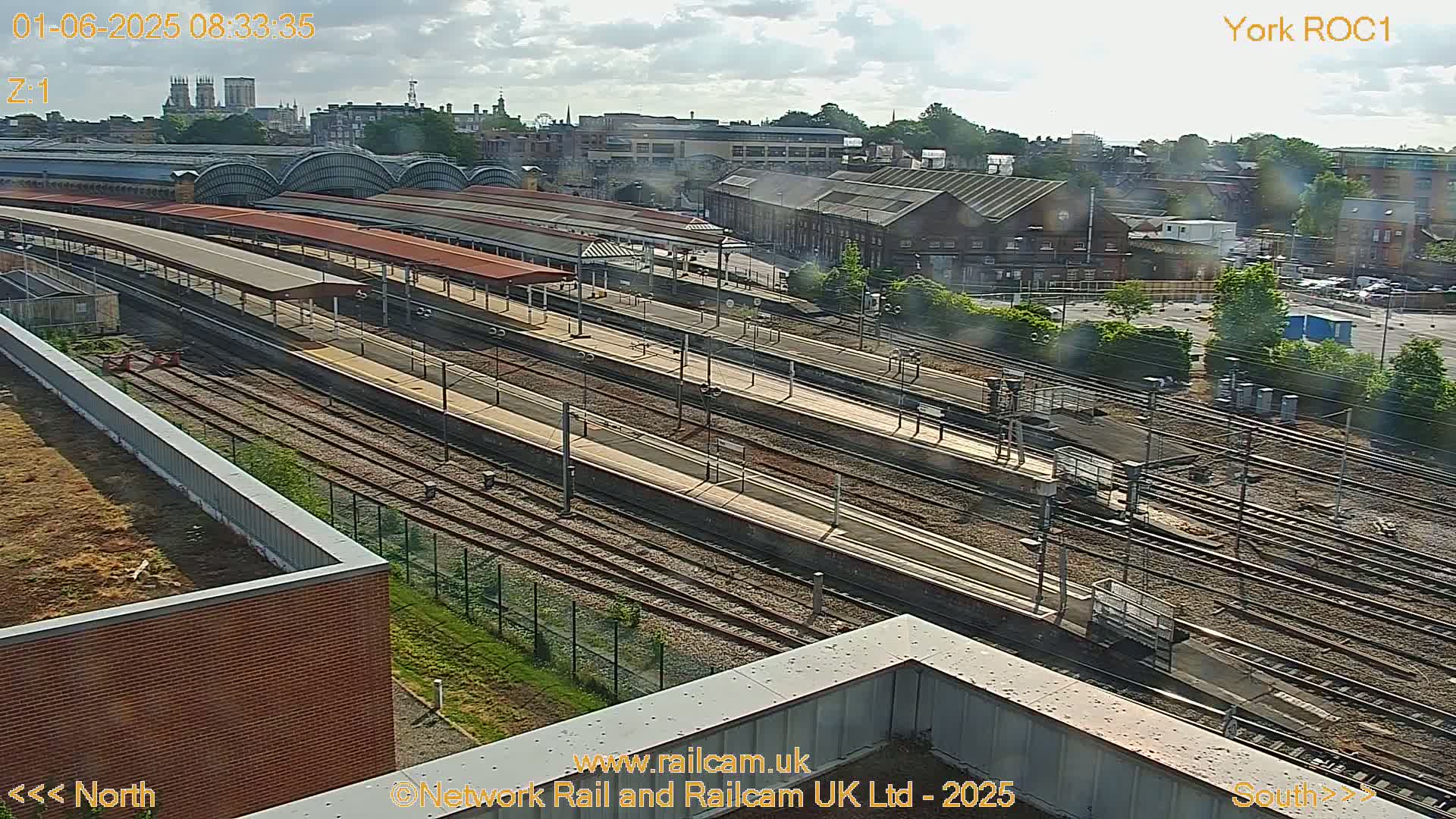 A high-angle view of a train station on a partly cloudy day, showing numerous train tracks and platforms extending into the distance, with a city skyline visible in the background.