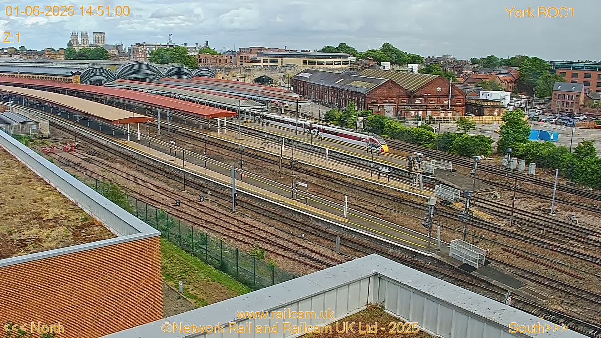 A high-angle view of a train station on an overcast day, showing multiple train tracks, platforms with covered waiting areas, and a train departing.