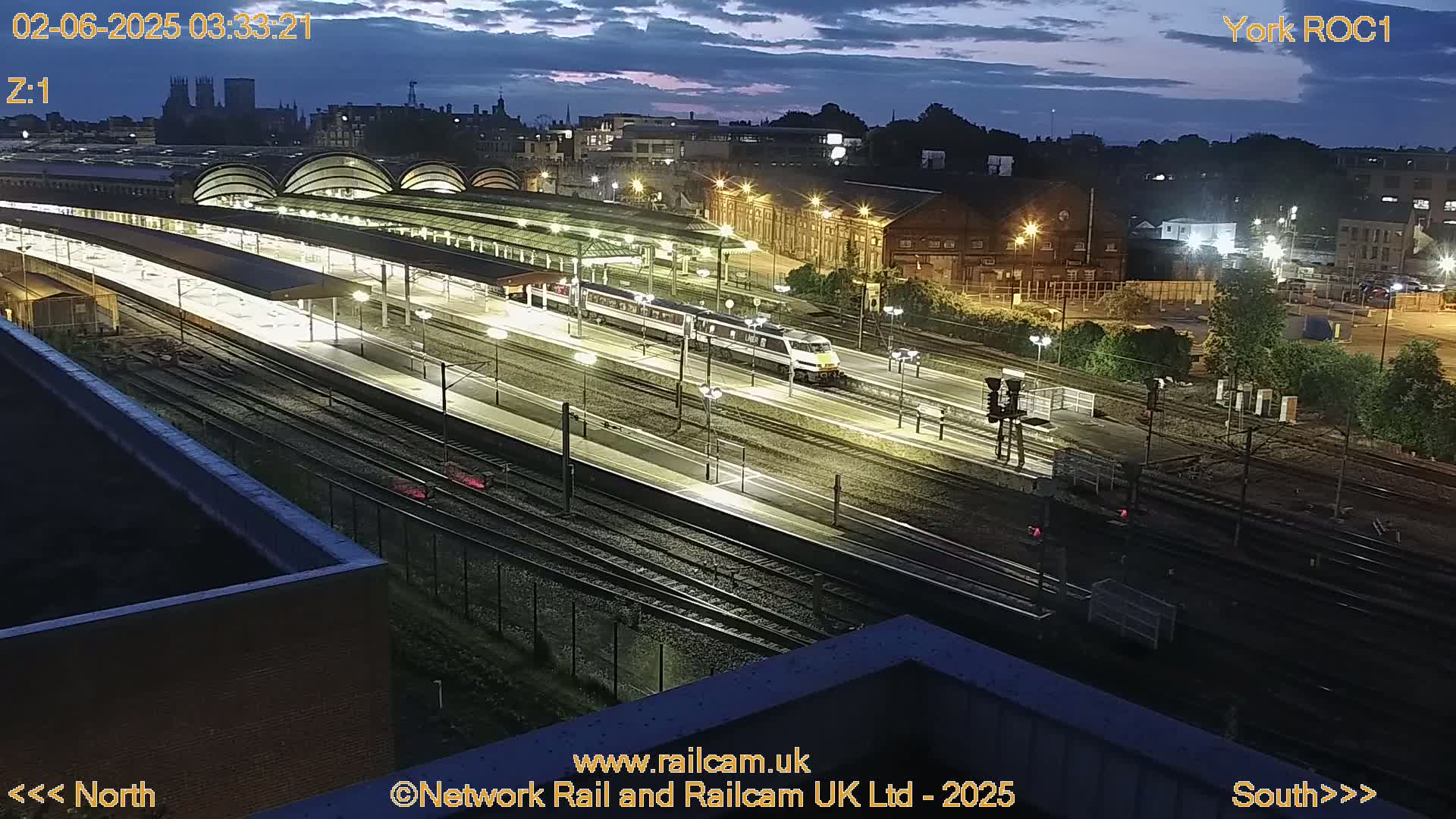 A train passes through a brightly lit train station at night under a twilight sky.