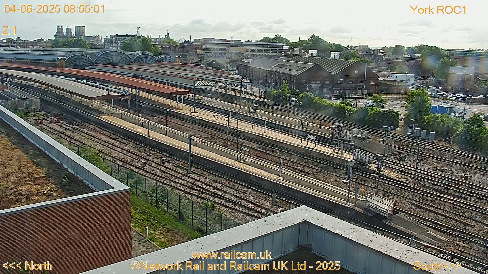A high-angle, long shot of a train station on a partly cloudy day, showing numerous train tracks, platforms, and surrounding buildings.