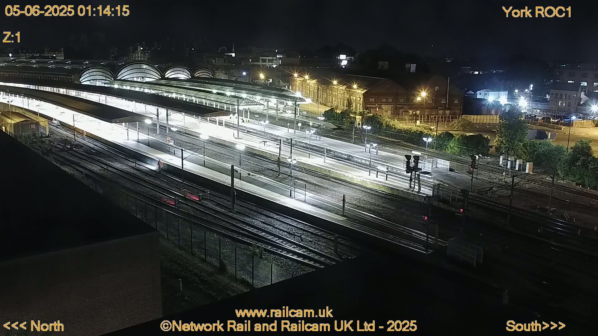 A nighttime, high-angle view of a brightly lit train station with multiple tracks and platforms.