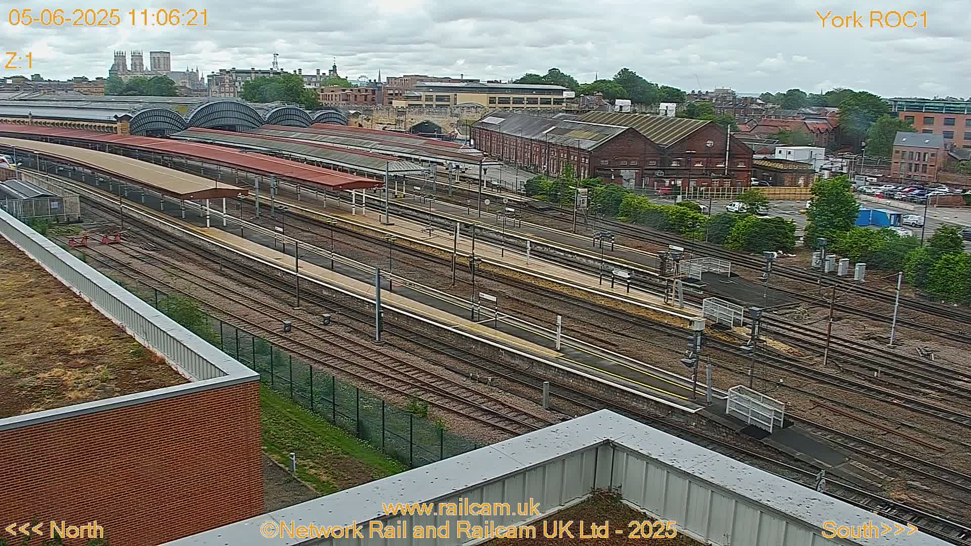 A high-angle, long shot of a train station on an overcast day, showing numerous train tracks and platforms, surrounded by buildings and parking lots.