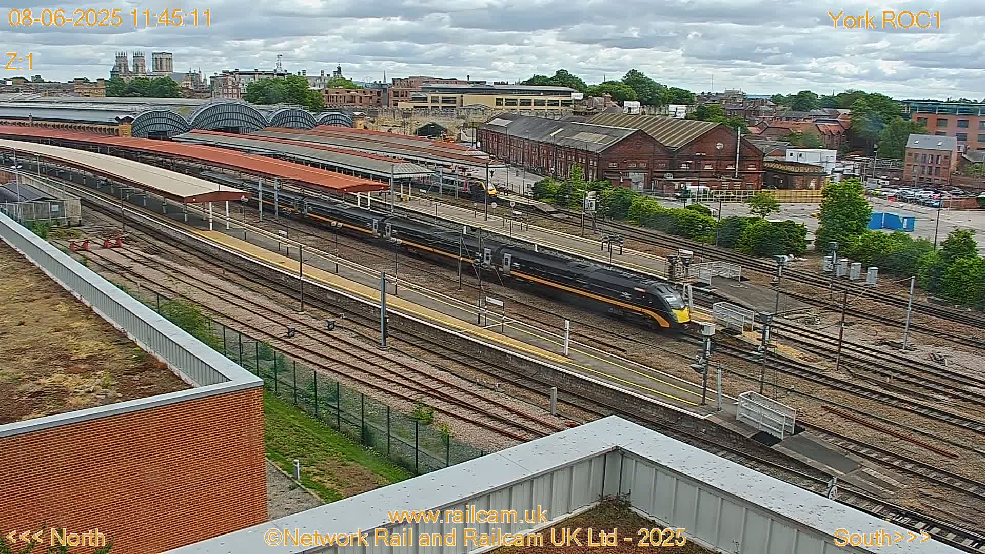 A high-angle view of a train station on an overcast day, showing multiple train tracks, platforms, and buildings, with two long black and yellow trains on the tracks.
