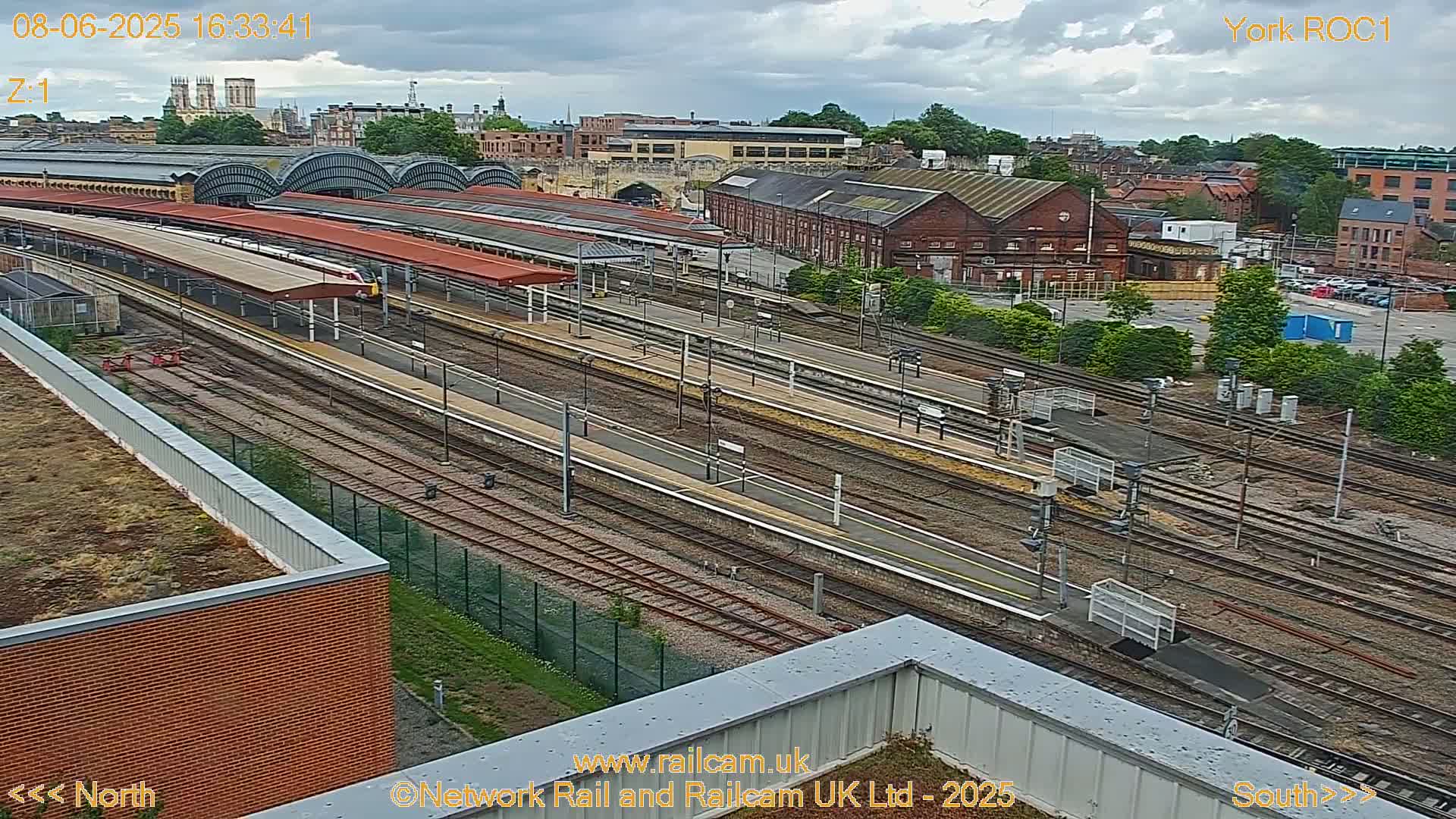 A high-angle view of a train station on an overcast day, showing multiple train tracks, platforms with covered waiting areas, and surrounding buildings.