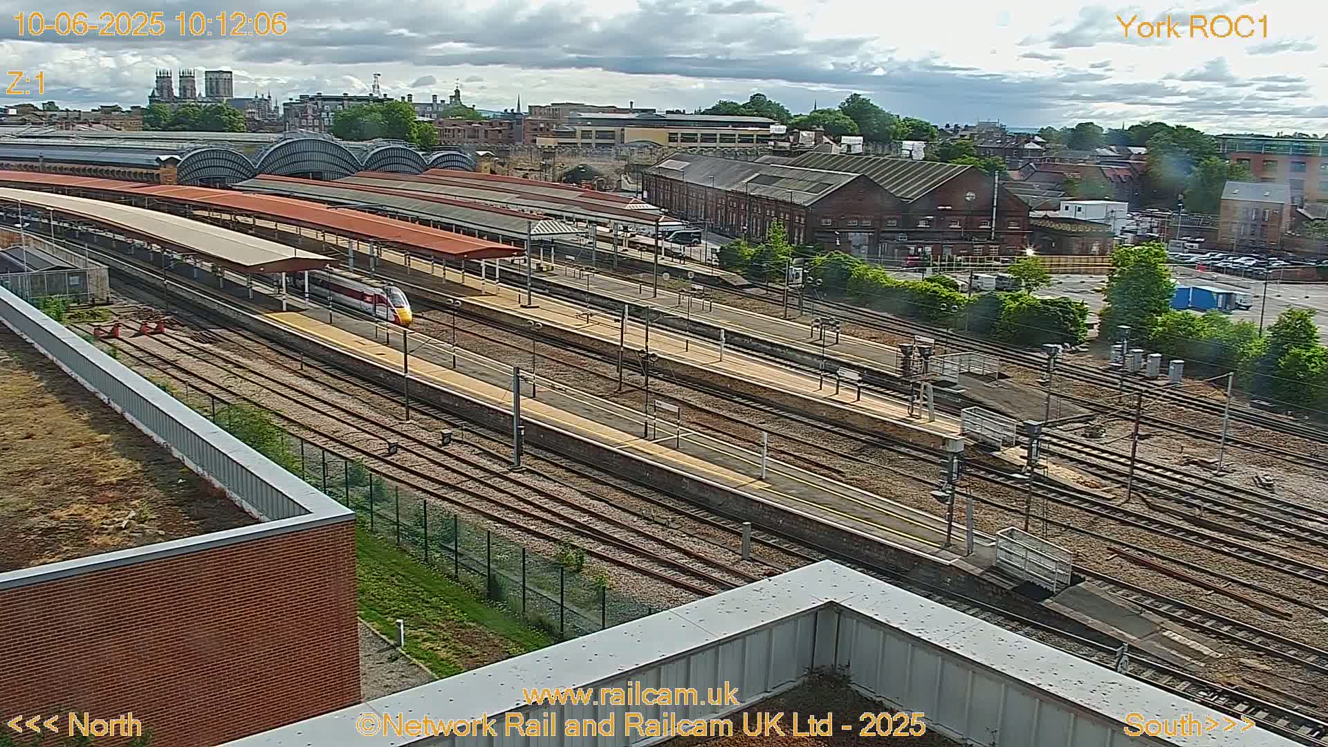 A high-angle view of a train station on a partly cloudy day, showing multiple train tracks, platforms with canopies, and a high-speed train departing.
