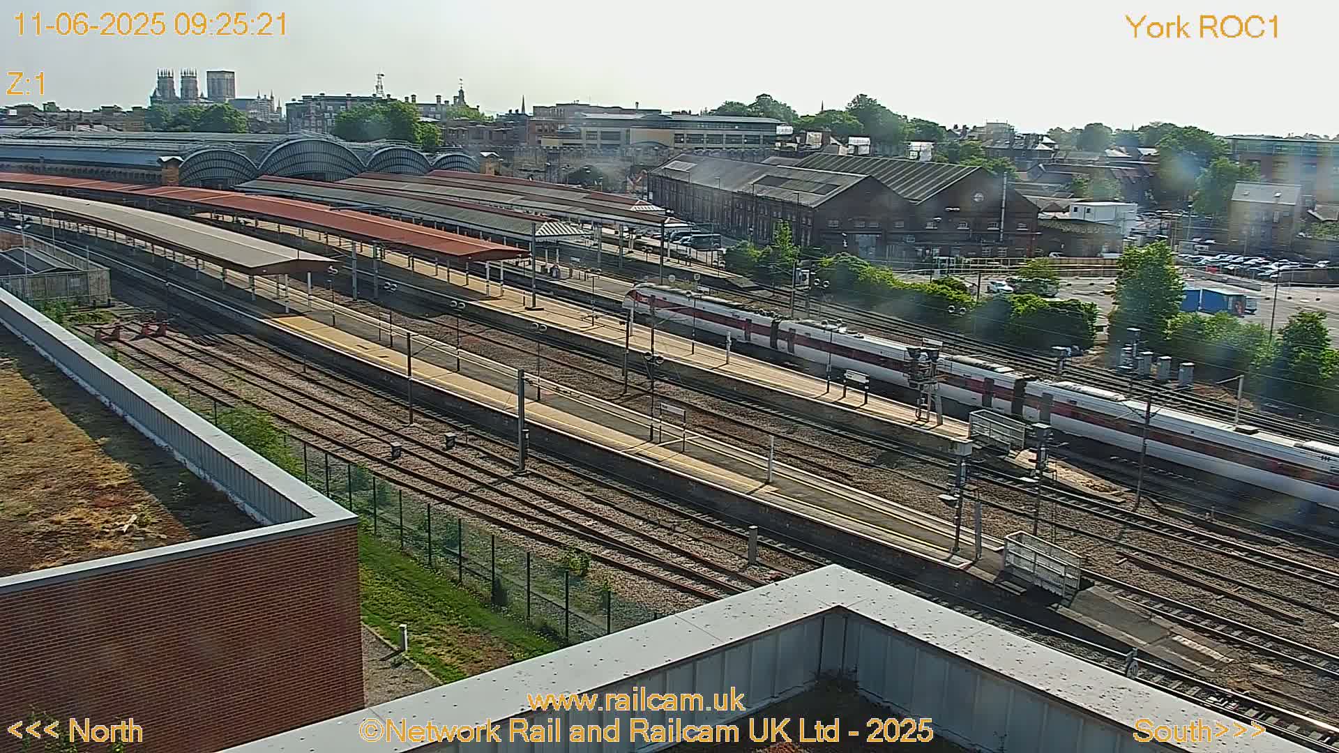 A high-angle view of a train station on a sunny day, showing multiple train tracks, platforms with covered waiting areas, and two trains alongside the platforms.