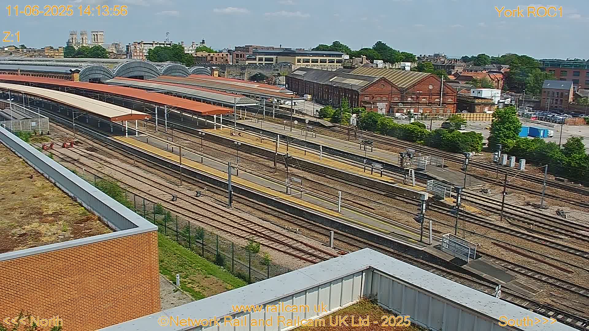 A high-angle, sunny view of a large railway station with numerous tracks and platforms, surrounded by industrial buildings and greenery.