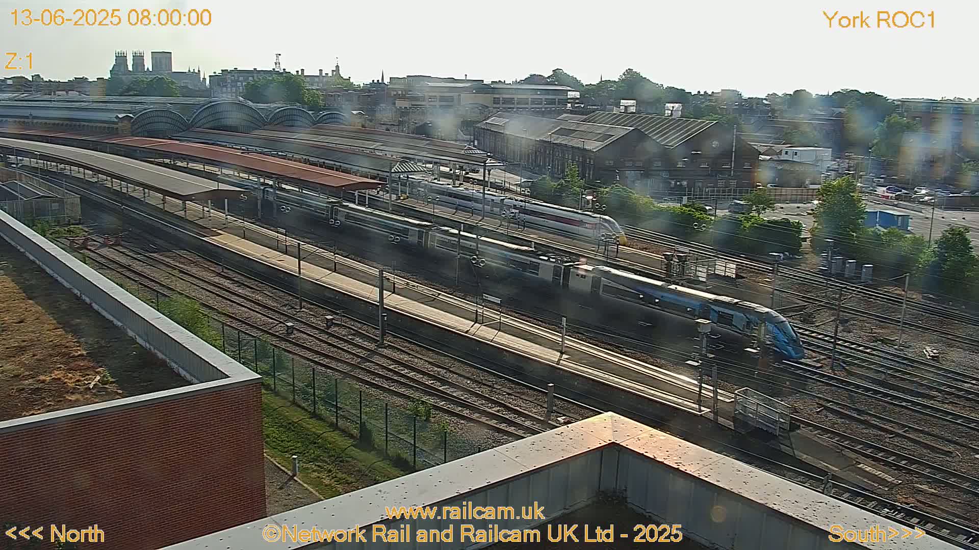 A high-angle, long shot of a train station on a sunny day, showing several trains on the tracks and platforms, with the city visible in the background.