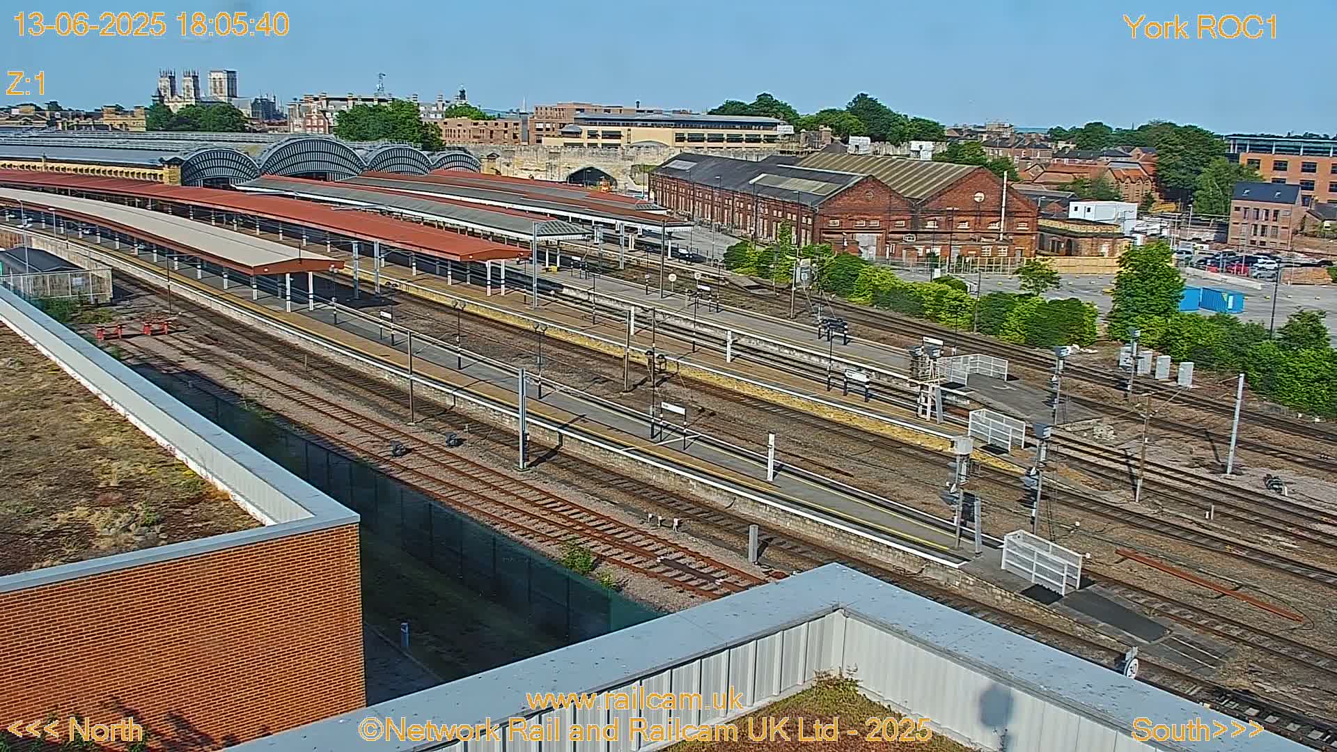A high-angle, sunny view of a train station with multiple tracks and platforms, surrounded by brick buildings and greenery.