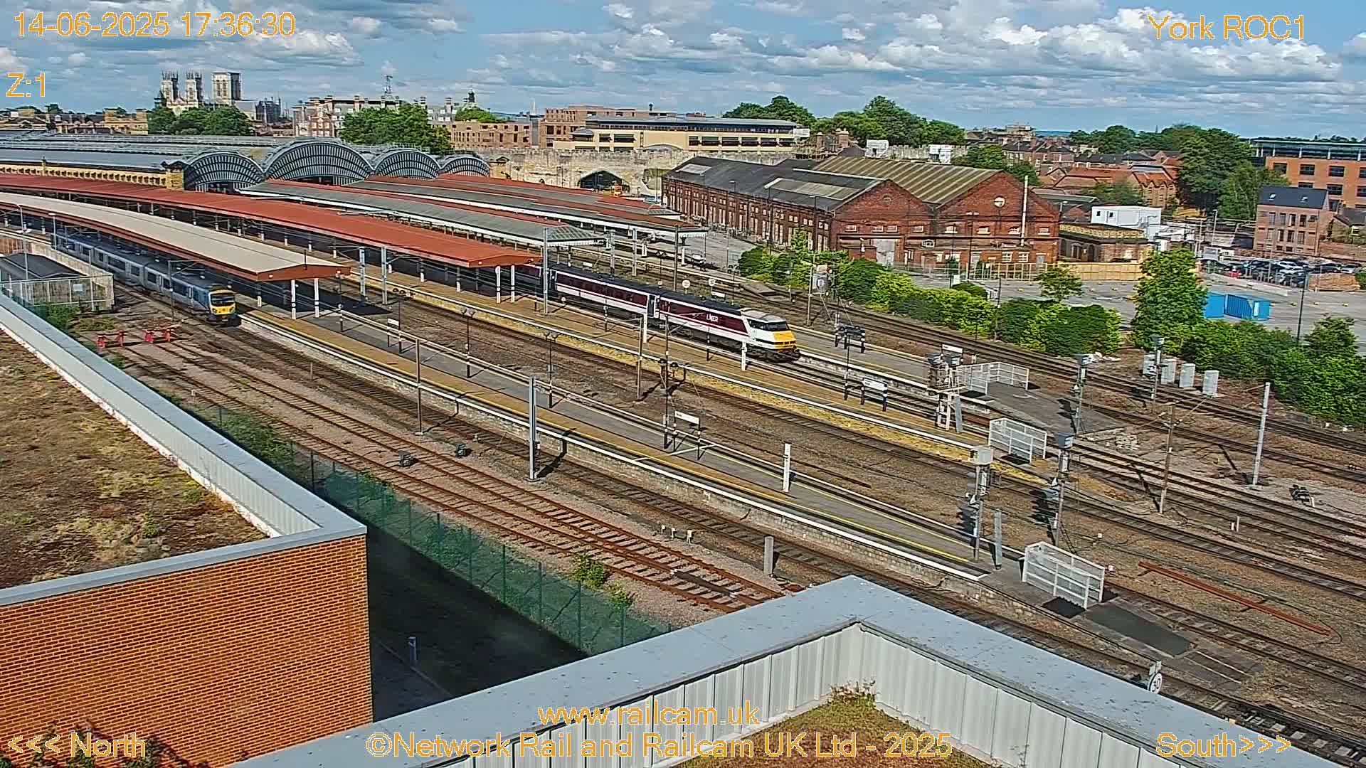 A high-angle view of a train station on a partly cloudy day, showing several train tracks, platforms with covered waiting areas, and multiple trains.