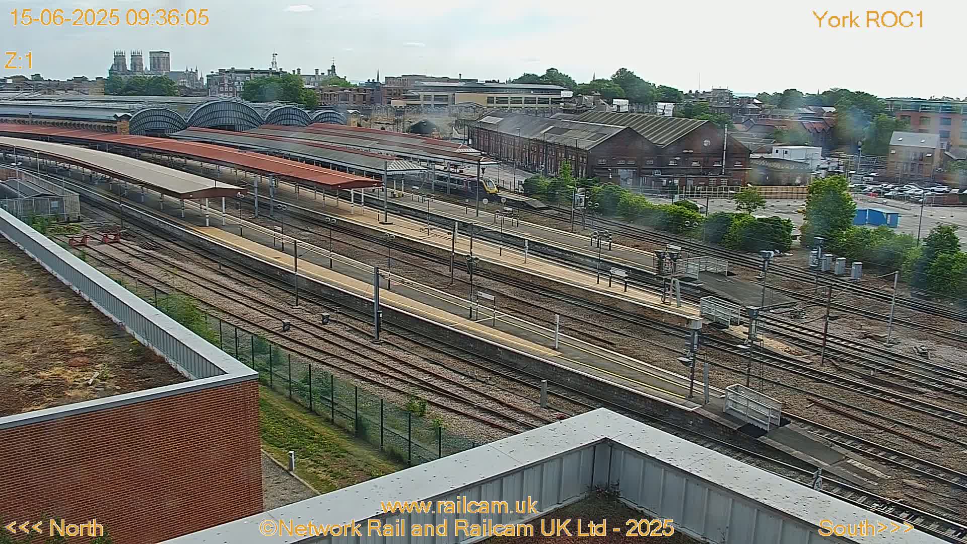 A high-angle view of a train station on a partly cloudy day shows numerous train tracks, platforms with canopies, and surrounding buildings.