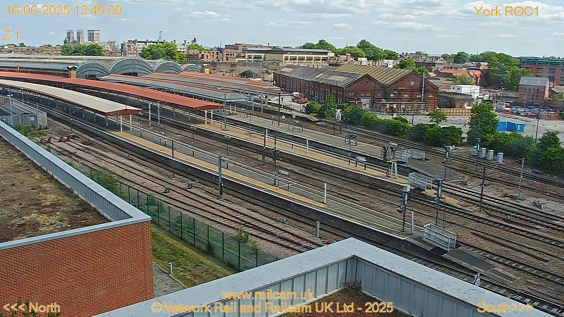 A high-angle view of a train station on a partly cloudy day, showing numerous train tracks and platforms extending into the distance, surrounded by buildings and greenery.