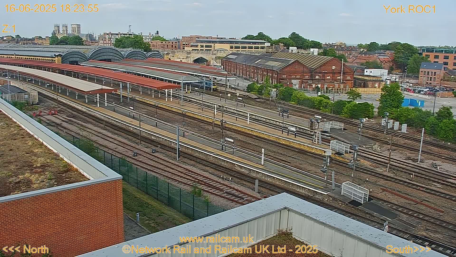 A high-angle view of a train station on a partly cloudy day, showing multiple train tracks and platforms, with surrounding buildings and greenery.