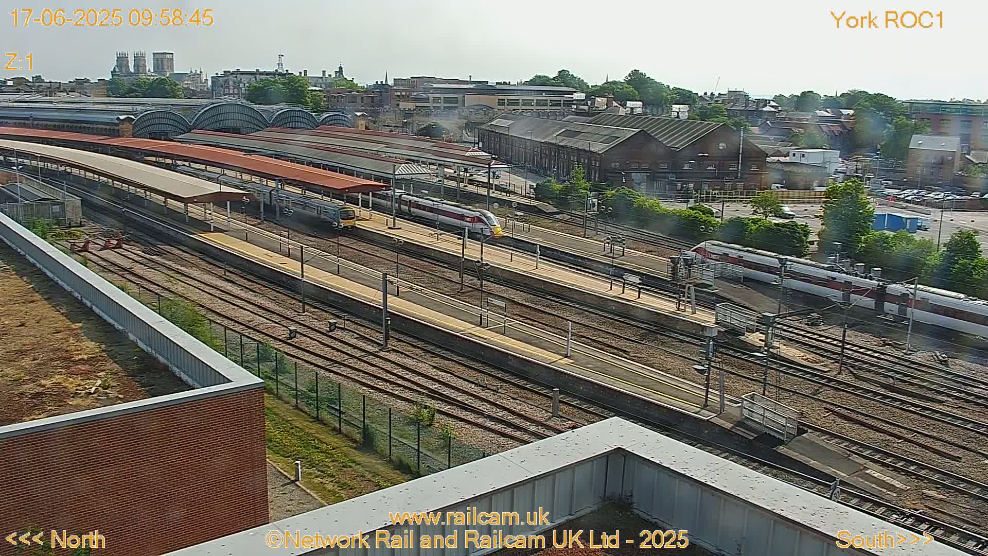 A high-angle view of a train station on a partly sunny day, showing multiple train tracks with several trains, and a city skyline in the background.