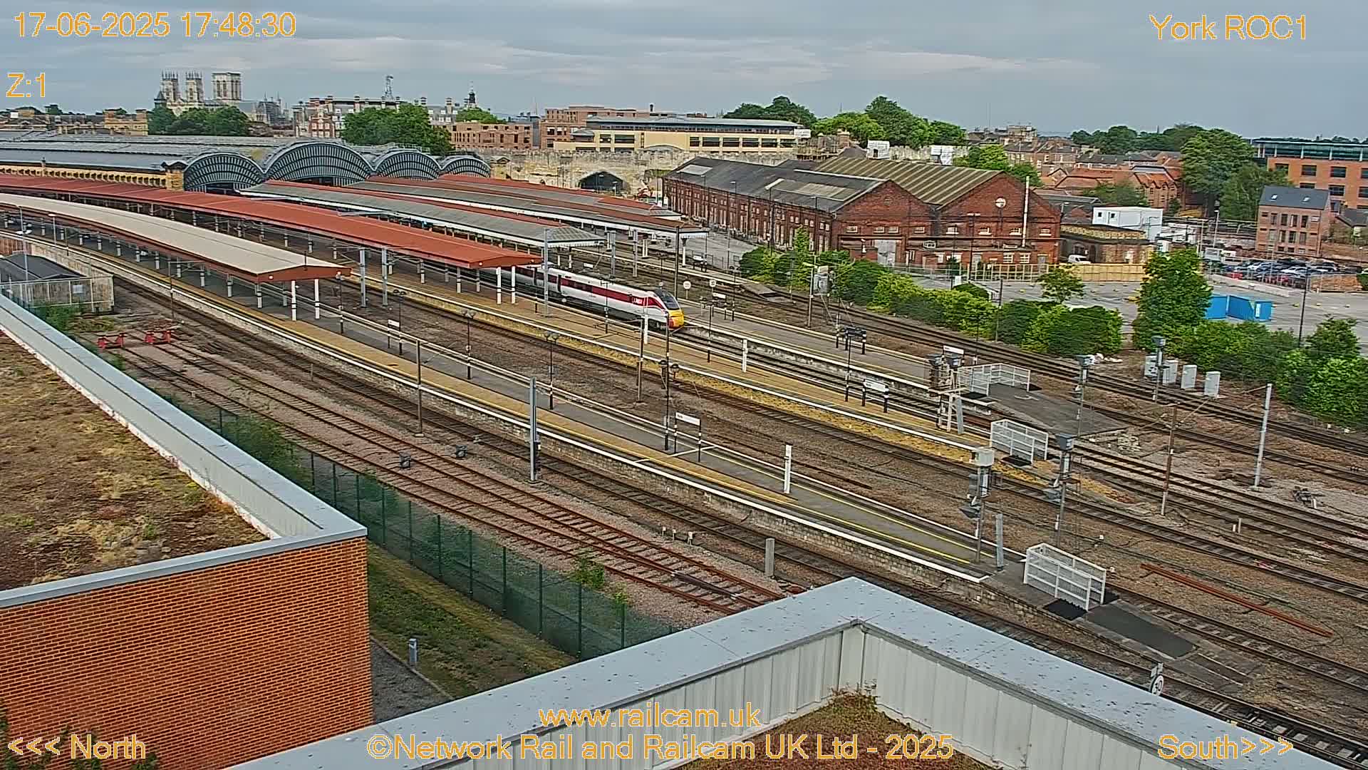 A high-angle view of a train station on an overcast day, showing a high-speed train passing through numerous tracks and platforms surrounded by buildings.