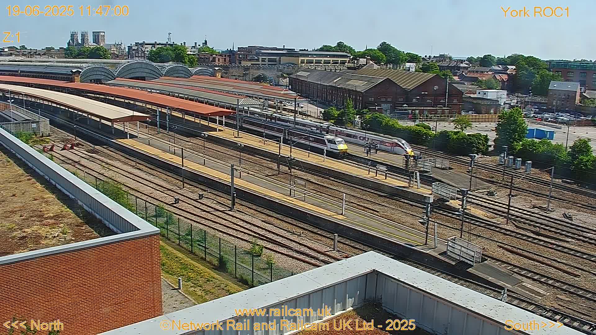 A high-angle view of a train station on a sunny day, showing multiple train tracks, platforms with covered waiting areas, and two high-speed trains.