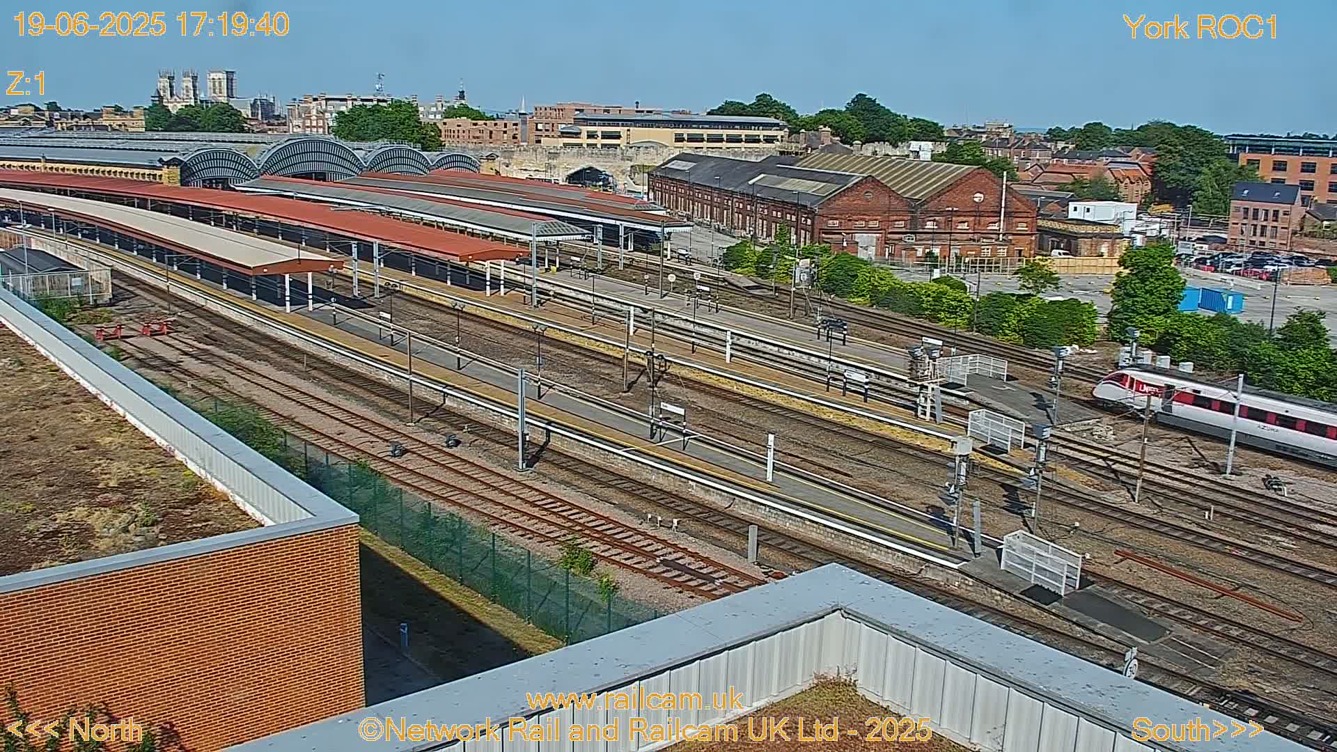 A high-angle view of a train station on a sunny day, showing multiple train tracks, platforms, and a train passing by.