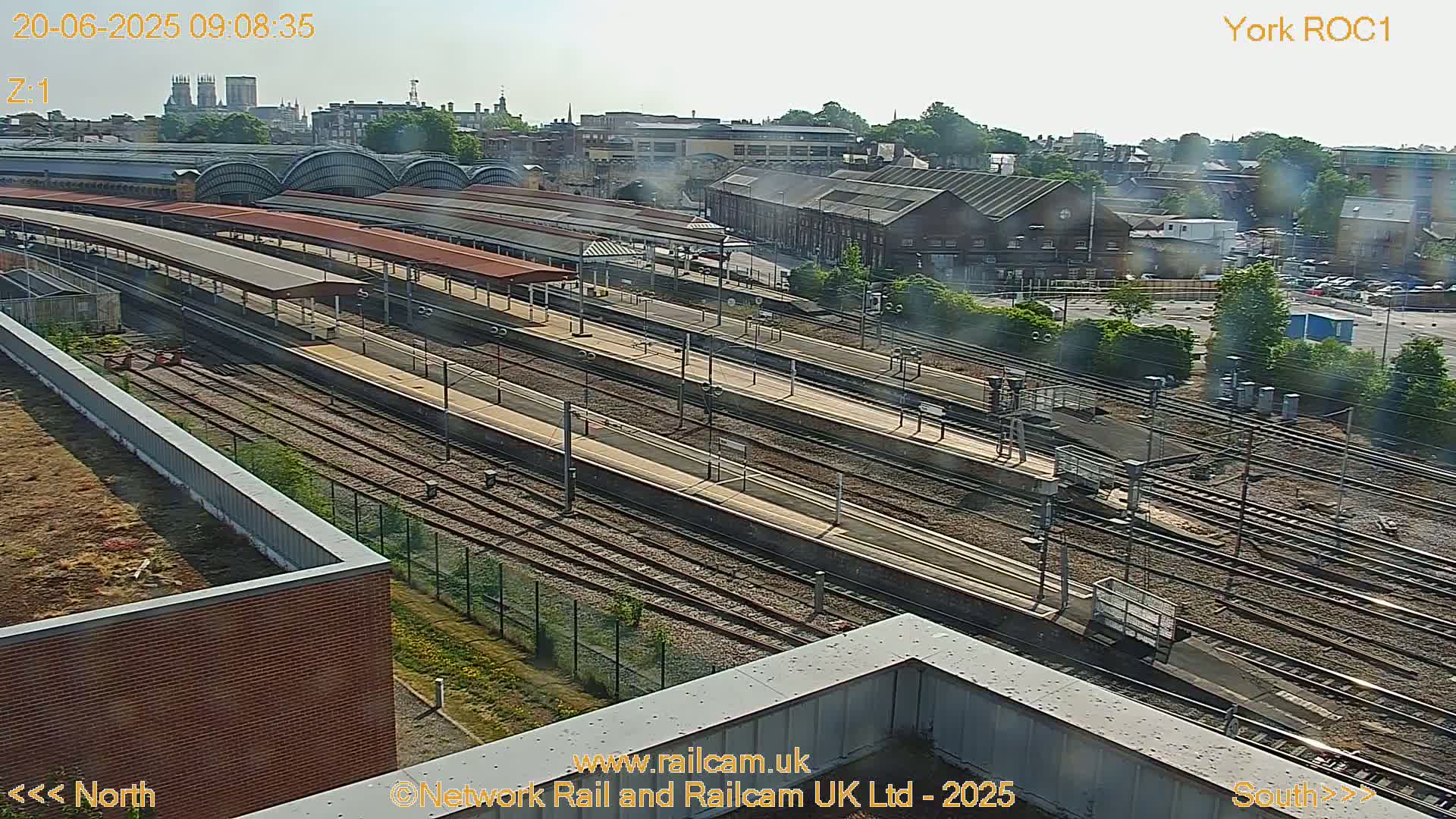 A high-angle view of a train station on a sunny day, showing numerous train tracks and platforms with surrounding buildings and vegetation.