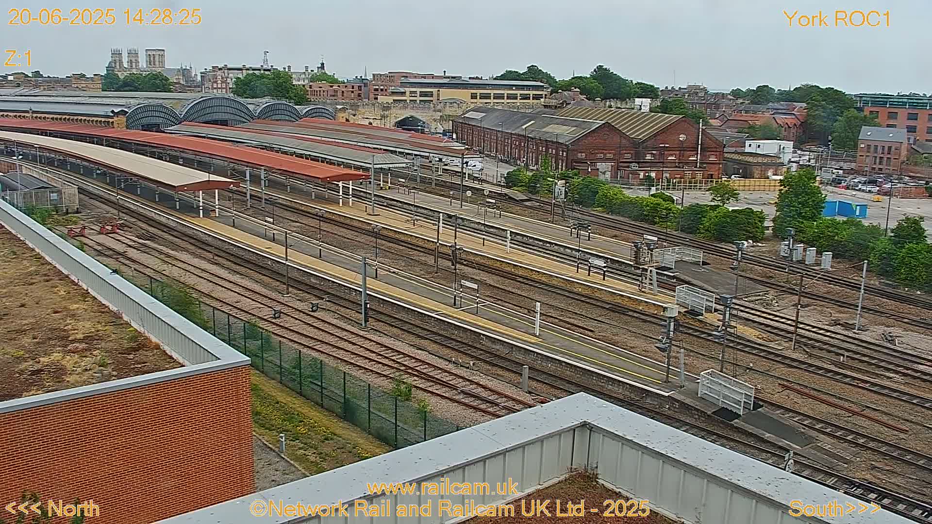 An aerial view of a train station on an overcast day, showing numerous train tracks, platforms with covered waiting areas, and surrounding buildings.