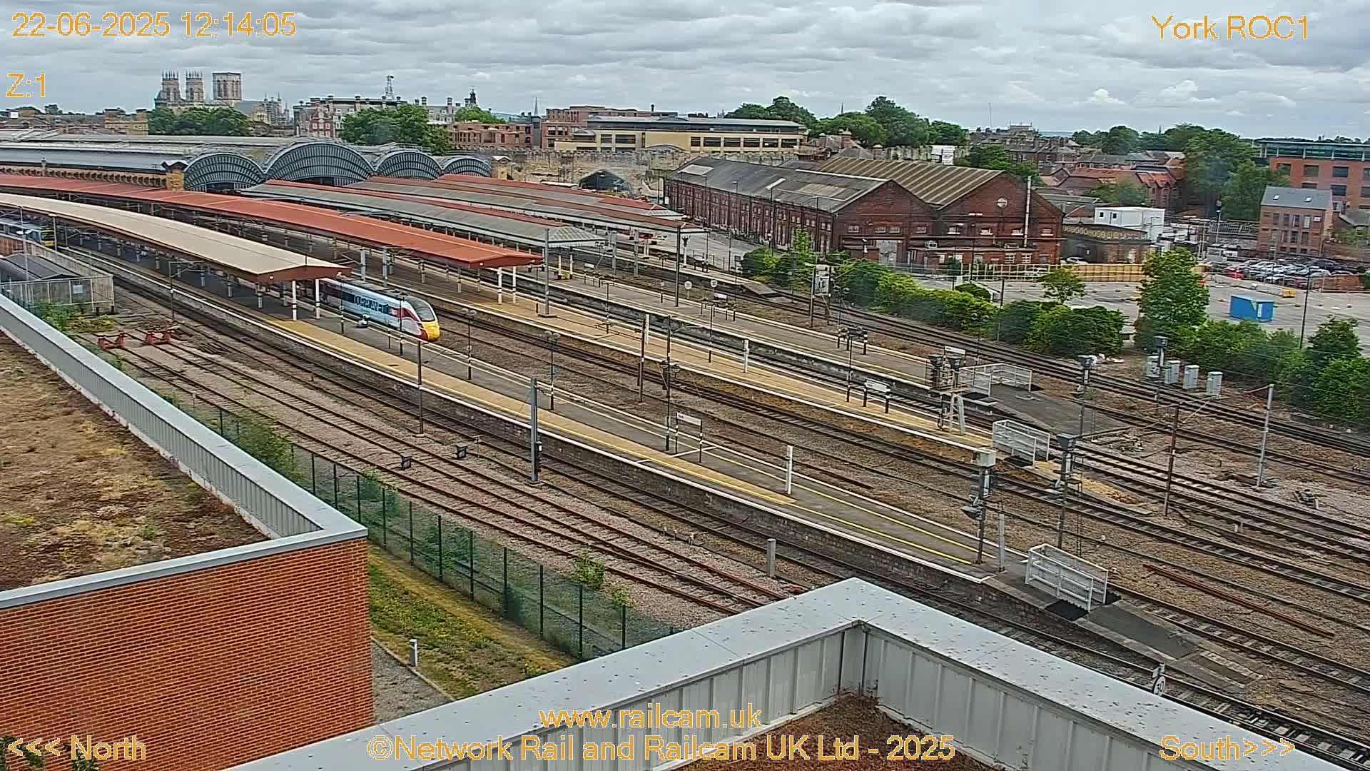 A high-angle view shows a train station with many tracks and a yellow and white high-speed train passing through under a partly cloudy sky.