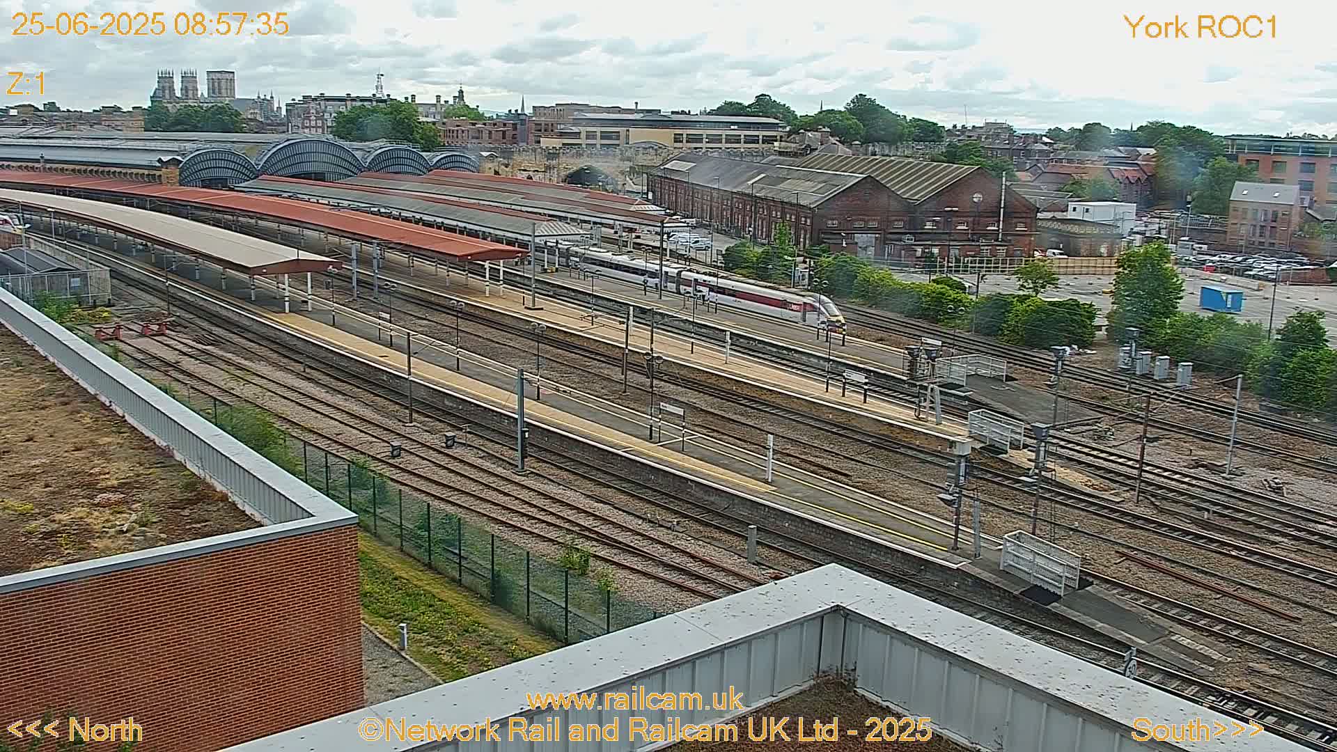 A high-angle view of a train station on a partly cloudy day, showing multiple train tracks, platforms with covered waiting areas, and a train passing through.