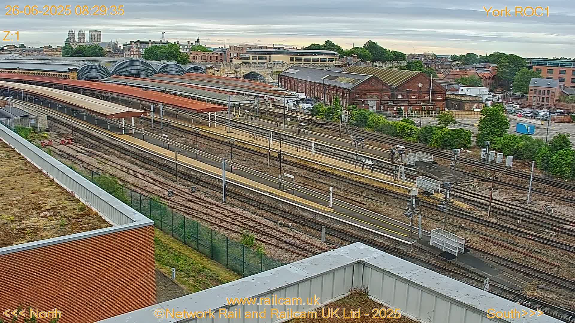 An aerial view of a train station and its surrounding area on an overcast day, showing numerous train tracks and platforms extending towards a cityscape in the distance.