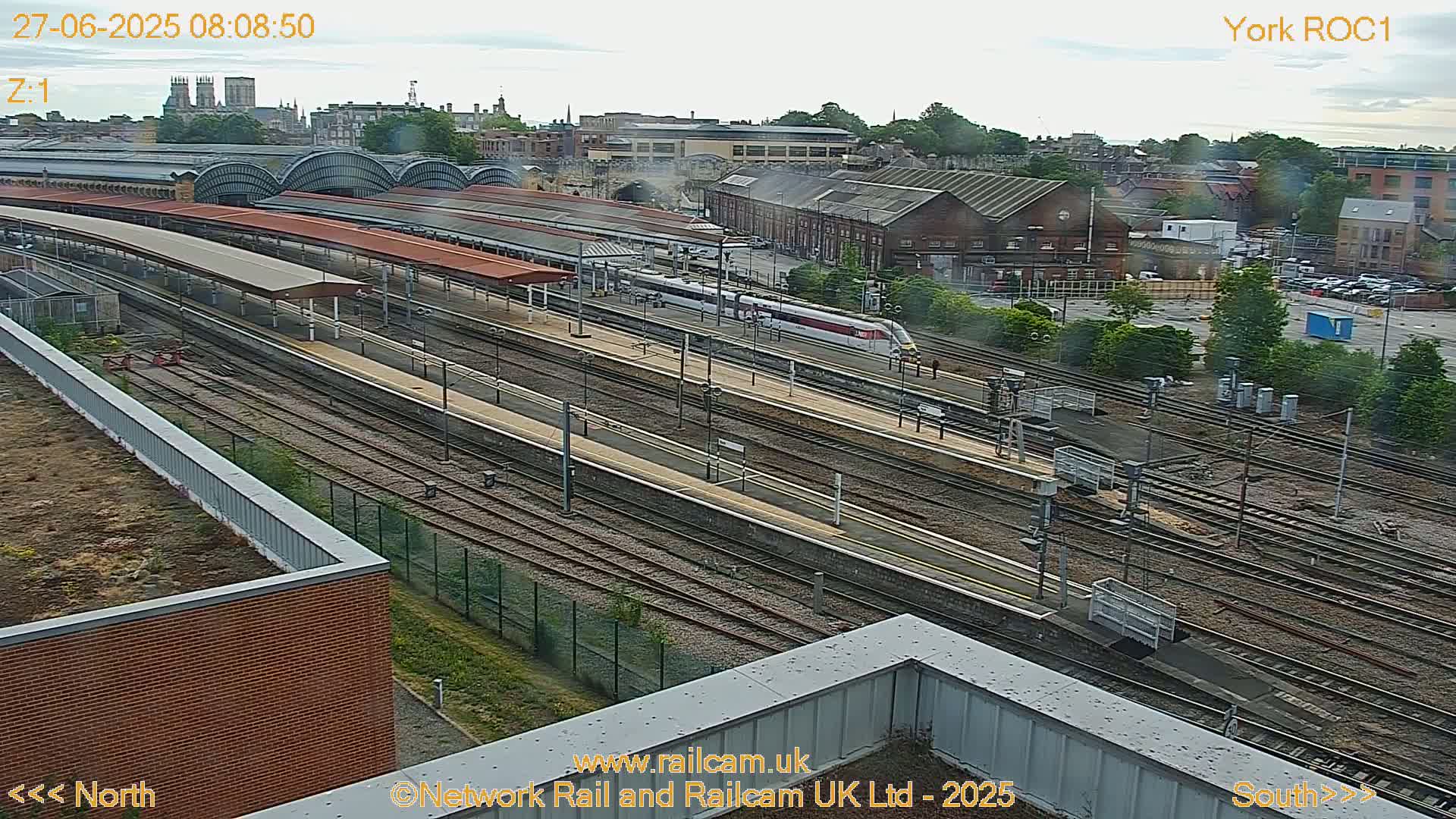 A high-angle, long shot shows a train station on an overcast day, with a train passing through numerous railway tracks.