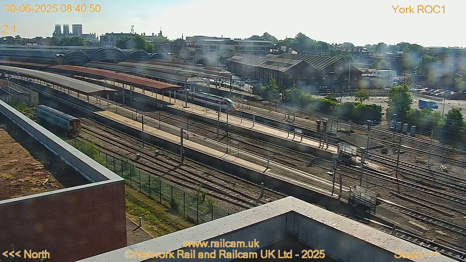 A high-angle view of a train station on a sunny day, showing multiple train tracks, platforms with trains, and surrounding buildings.
