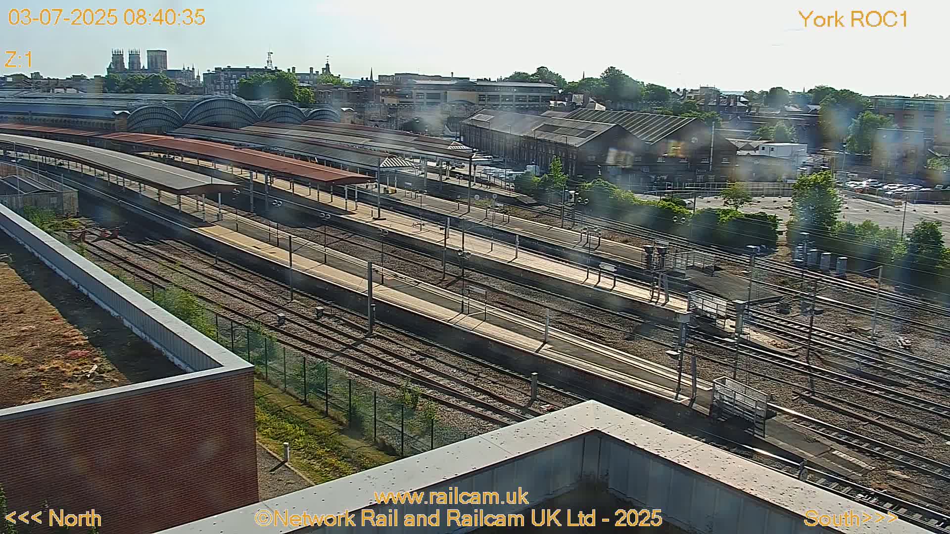 A high-angle, long shot of a train station on a sunny day, showing numerous train tracks, platforms, and surrounding buildings.