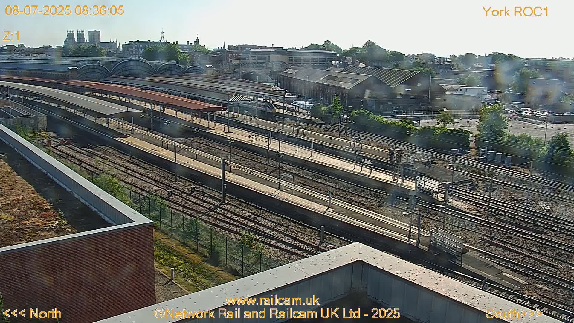 A high-angle, long shot of a train station on a sunny day, showing numerous train tracks and platforms.