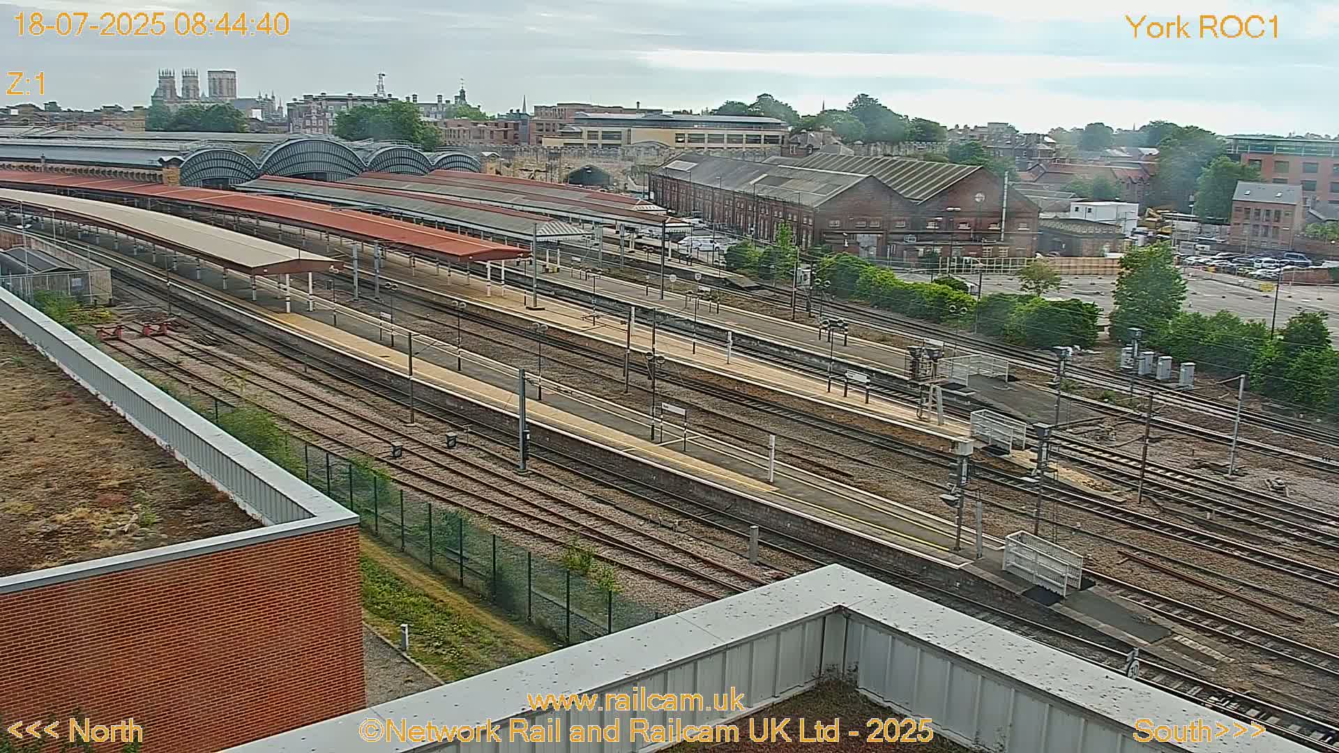 A high-angle view of a train station's expansive rail yard on an overcast day, with numerous tracks and platforms visible, and buildings and a cityscape in the background.