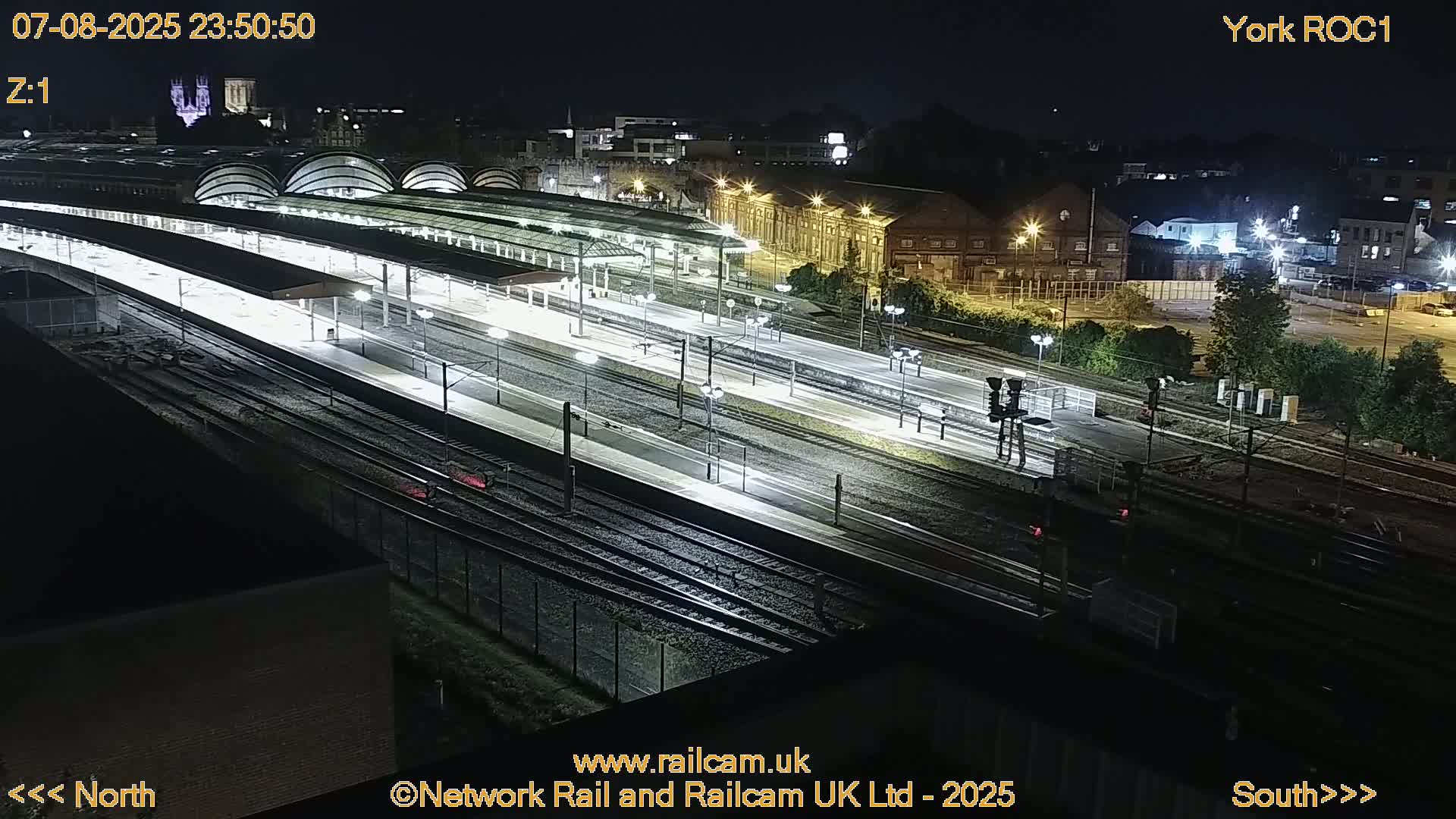 A nighttime, high-angle view of a brightly lit train station with multiple tracks and platforms.