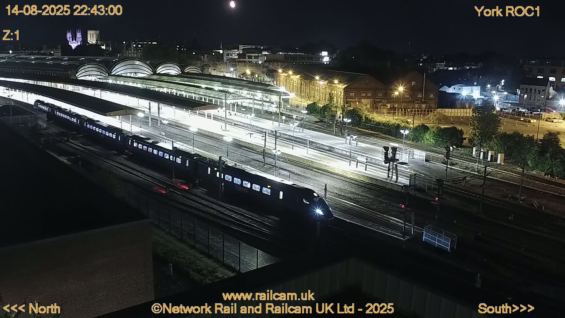 A long, dark train moves through a brightly lit train station at night.