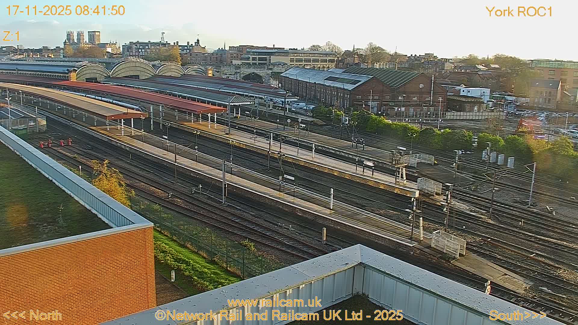 An expansive view of a large railway station with numerous tracks and platforms unfolds under a bright morning sky, bordered by a city skyline featuring prominent historical architecture.